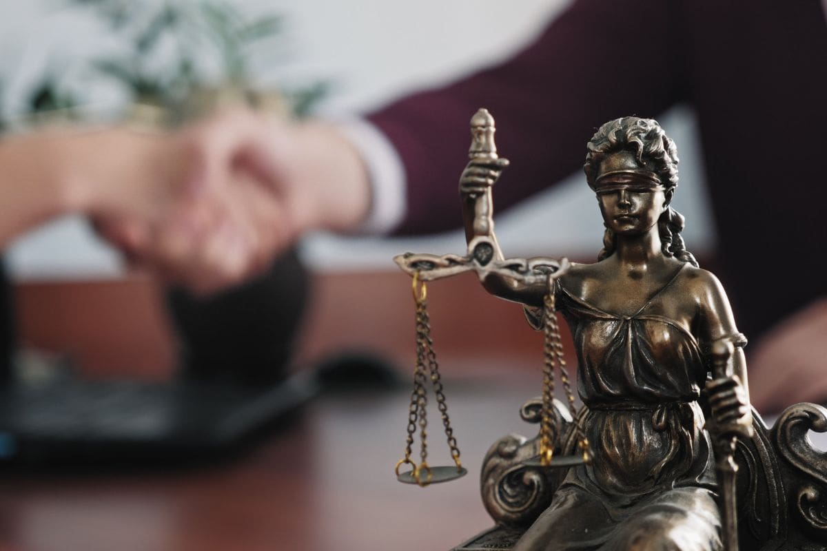 A lady justice statue with a person shaking hands in the background.